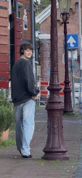 A young person wearing headphones and light-wash jeans smiles while looking back over their shoulder on an urban street